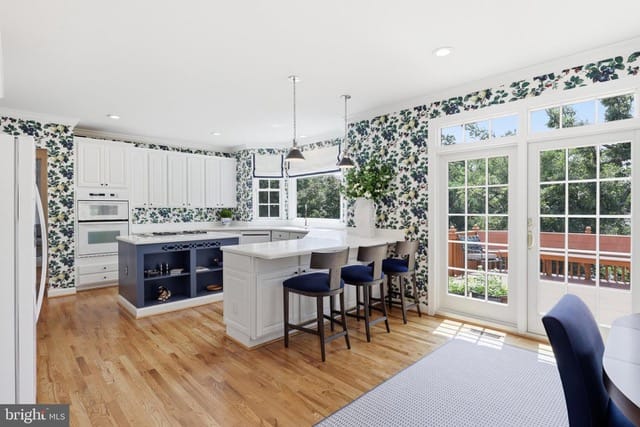 Bright open kitchen with white cabinets, a blue island, hardwood floors, floral wallpaper, and French doors opening to a deck.