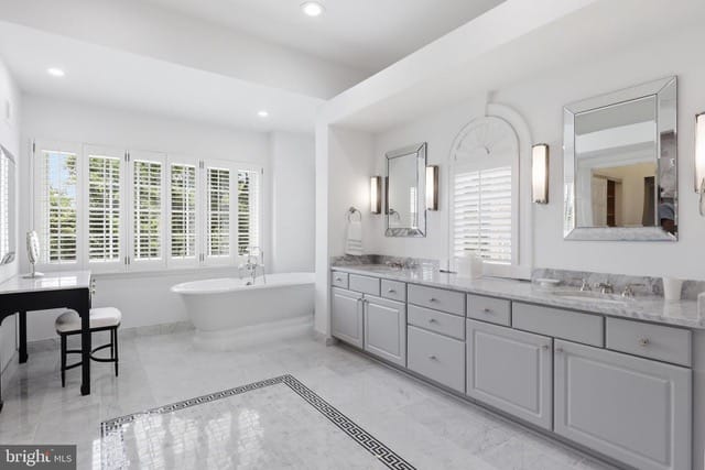 Bright white bathroom featuring a double-sink vanity with marble countertops, a freestanding tub by a window with shutters, and a tiled floor.