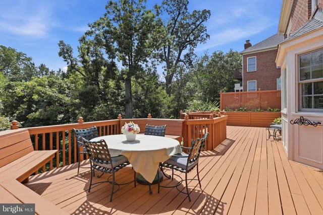 Wooden deck with a round table and four chairs, a beige tablecloth, a flower centerpiece, railing, and trees against a blue sky.