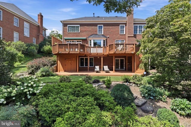 Two-story brick house with a large wooden deck overlooking a lush landscaped backyard garden.