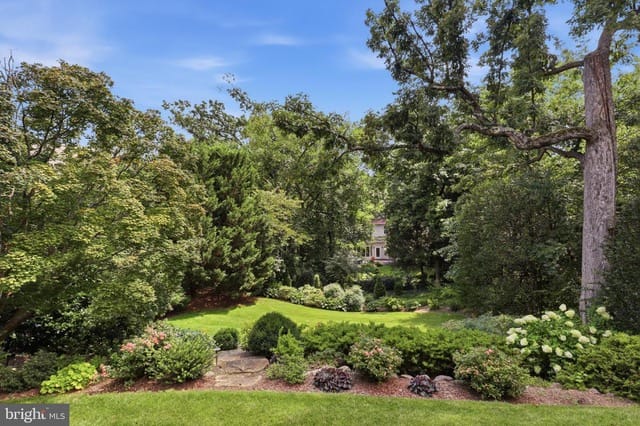 A manicured garden with green lawn, trimmed shrubs, and tall trees, with a path in the foreground and a house visible in the distance.
