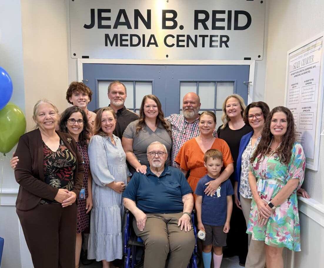 Group photo in the Jean B. Reid Media Center lobby with a man in a wheelchair at center and smiling staff and visitors surrounding him.