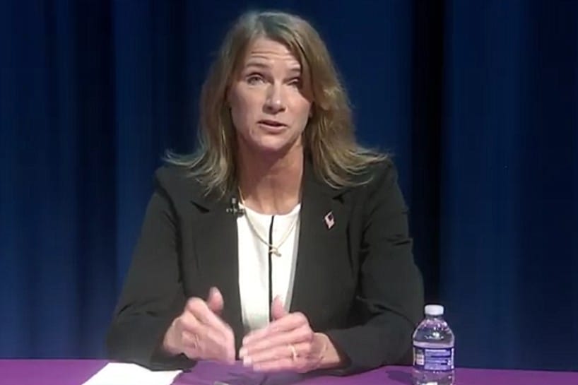 Woman in a blazer speaks at a table, with a water bottle on the purple tablecloth and a microphone clipped to her lapel.