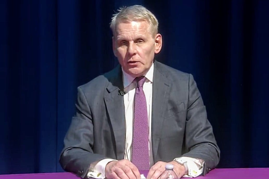 Man in a gray suit and pink tie sits at a purple table with a water bottle, speaking on stage behind a dark blue curtain.