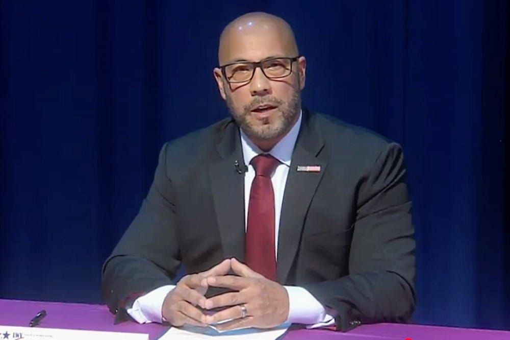 Bald man in a dark suit and red tie speaks at a panel, hands clasped on a purple table.