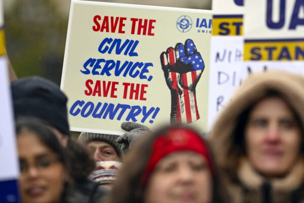 Protest sign reading 'Save the Civil Service, Save the Country!' with a blue-red fist in U.S. flag colors, surrounded by a crowded rally.