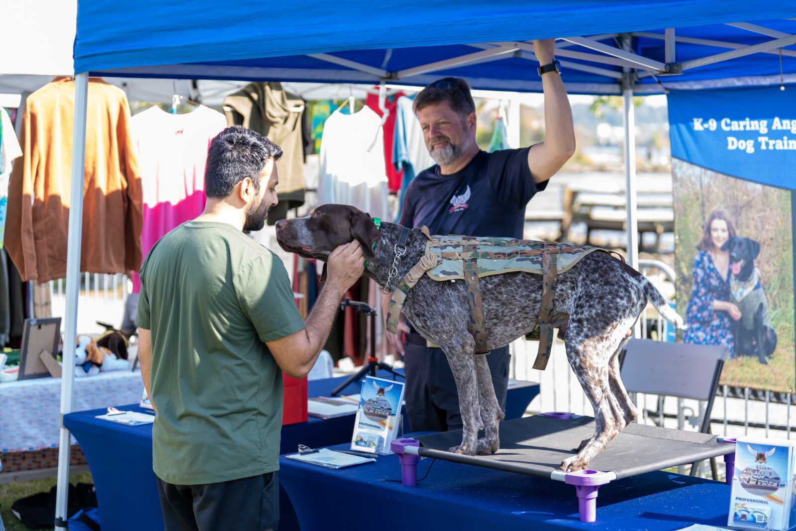 Man in green shirt pets a speckled grey hunting dog in a camouflage harness on a table at a K-9 training booth under a blue canopy.
