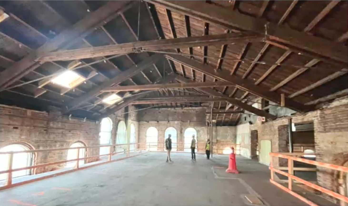 Interior of a brick warehouse with exposed wooden roof trusses and arched windows; four people stand near the far wall at center.