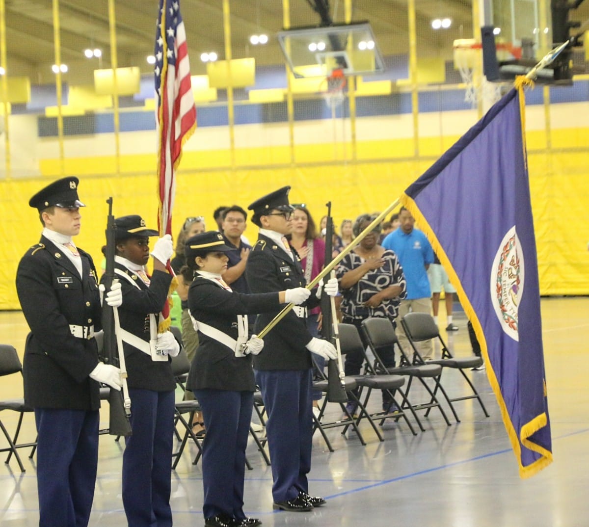 Color guard in dress uniforms standing at attention, holding the American flag and a blue ceremonial flag in a gymnasium ceremony.