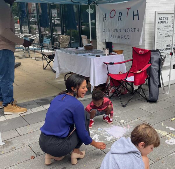 Woman crouching beside a child drawing with chalk on the sidewalk near a booth with a North Old Town Alliance banner.