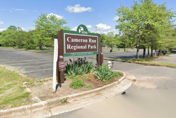 Cameron Run Regional Park sign in a landscaped roundabout with trees and a parking lot behind it, under a blue sky