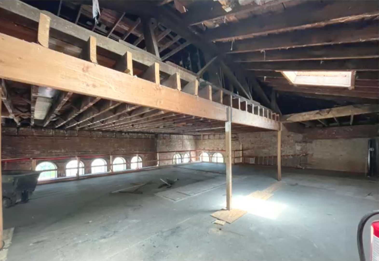 City Hall interior renovation (via City of Alexandria) | ALXnow Old brick loft with a wooden mezzanine, exposed ceiling beams, and arched windows along the wall; dusty concrete floor and scattered debris.