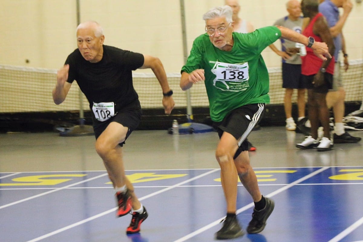 Two elderly men sprinting on an indoor track, bibs 108 and 138, leaning forward in a race.