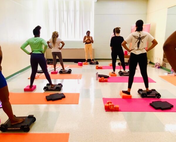 Instructor leads a group cardio class in a bright studio; participants stand on colorful yoga mats with mini step platforms nearby.