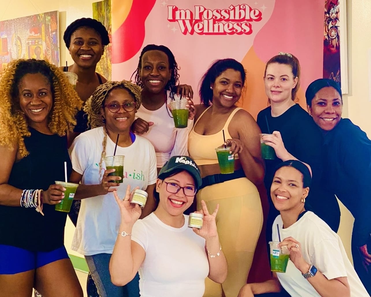 Grupo de nueve mujeres posando juntas en un evento de bienestar en interiores, sosteniendo bebidas verdes, frente a un fondo rosa de 'I'm Possible Wellness'.