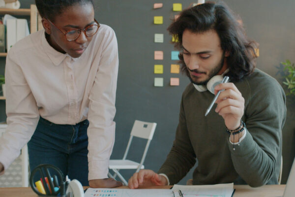 Two colleagues collaborate at a desk, examining papers as one points with a pen and the other looks on over the document.