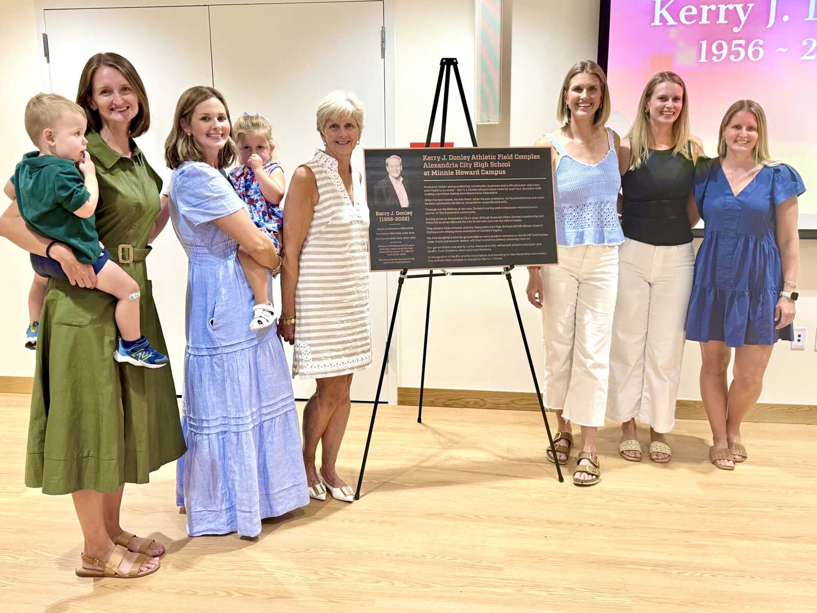 Group of adults (and two children) posing beside a commemorative plaque on a tripod easel in a bright room; plaque includes a portrait and text about Kerry J. Donley Athletic Field Complex at Alexandria City High School Minnie Howard Campus.