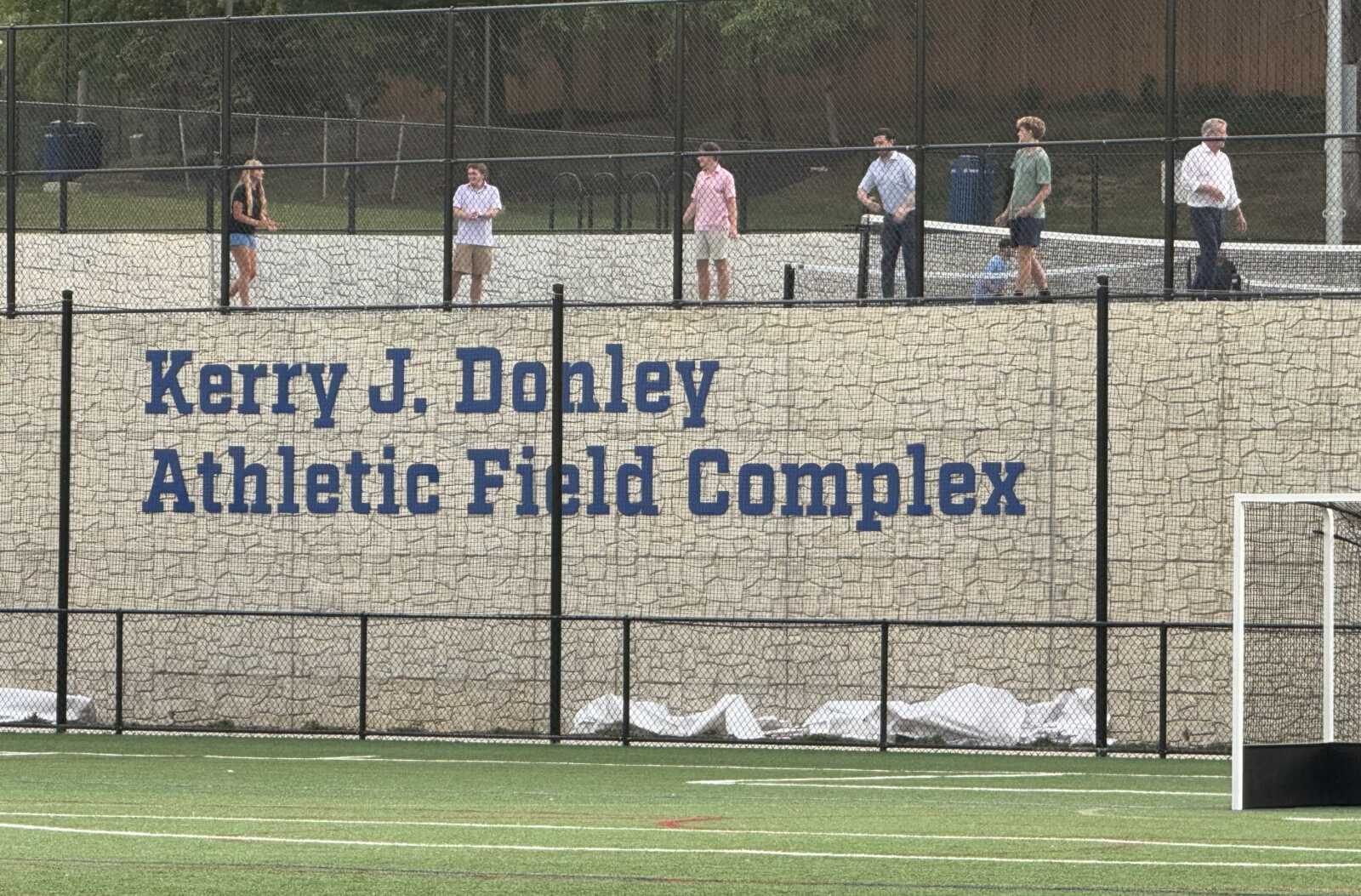 Brick wall with blue lettering: 'Kerry J. Donley Athletic Field Complex' behind a chain-link fence; people stand along the walkway above it, on a sports field.