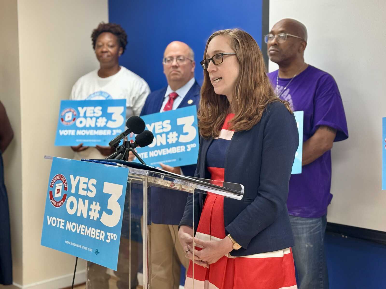 Woman speaking at a podium with campaign signs reading Yes on 3 in the background; supporters stand behind her.