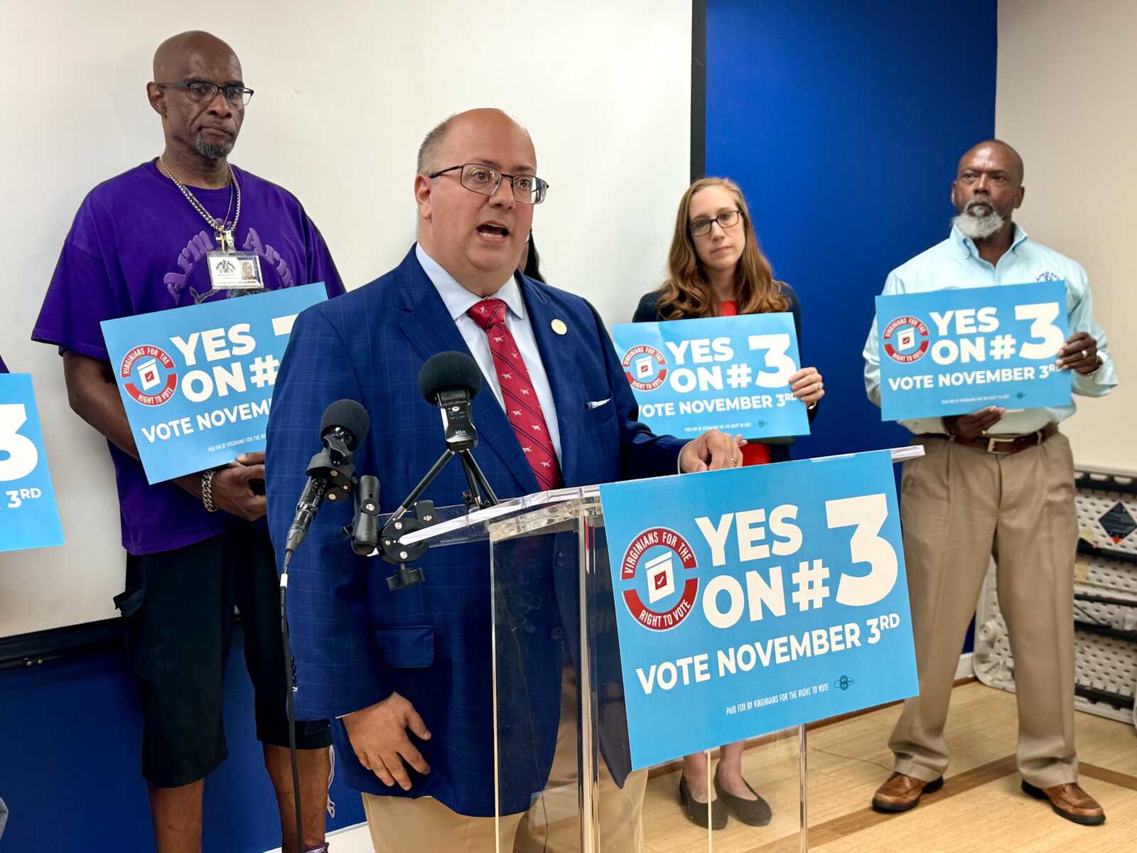 Speaker in a blue suit addresses a press conference at a clear podium with multiple microphones, surrounded by supporters holding Yes on 3 signs for voting on November 3.