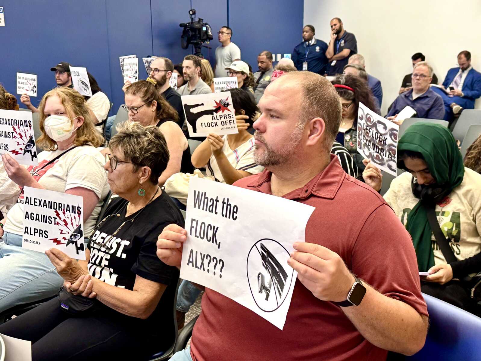 Group of diverse protesters seated in a room, holding signs opposing ALPRs with slogans like 'ALEXANDRIANS AGAINST ALPRS' while a camera stands on a tripod in the background.