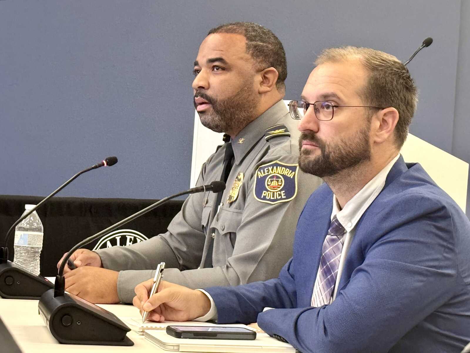 Two men at a panel discussion with microphones; a police officer in uniform and a suited man sit at a table.