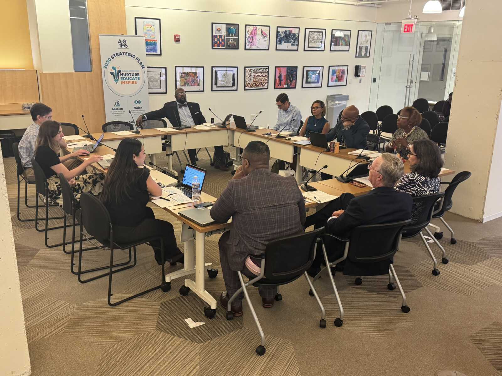 Group of professionals seated around a U-shaped conference table in a bright room, each with laptops and papers for a meeting, a banner reading '2030 Strategic Plan' visible in the background.
