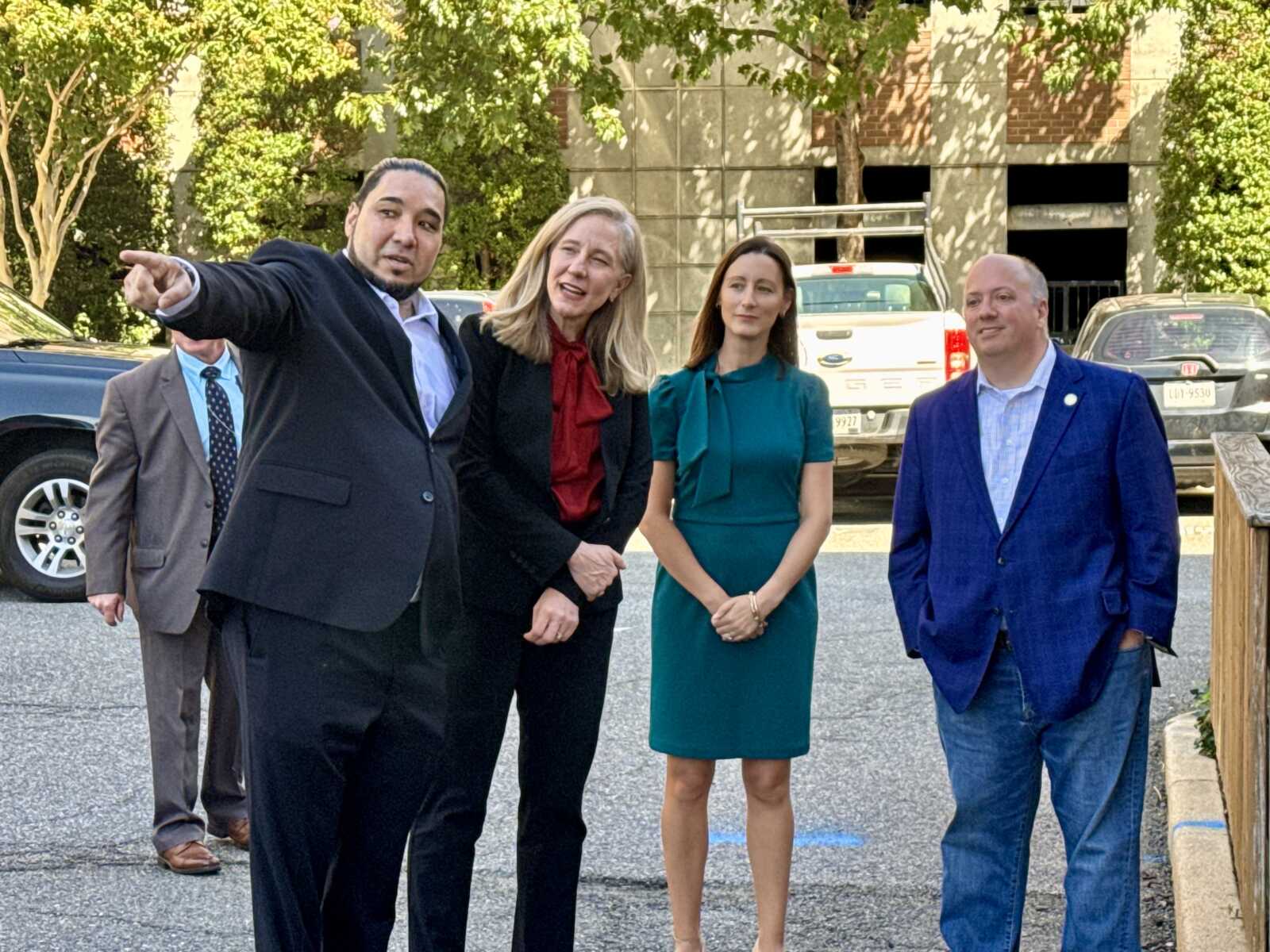 Man in a dark suit points to the left while four colleagues stand nearby—three women and a man, all in business attire, outdoors in a parking lot.