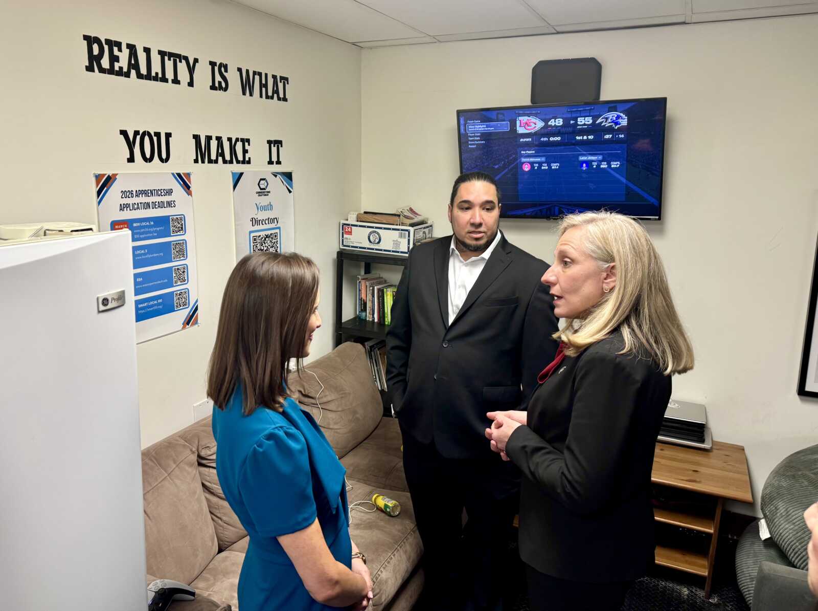 Three professionals in a small office discuss a topic, with a wall slogan reading 'Reality is what you make it' behind them.