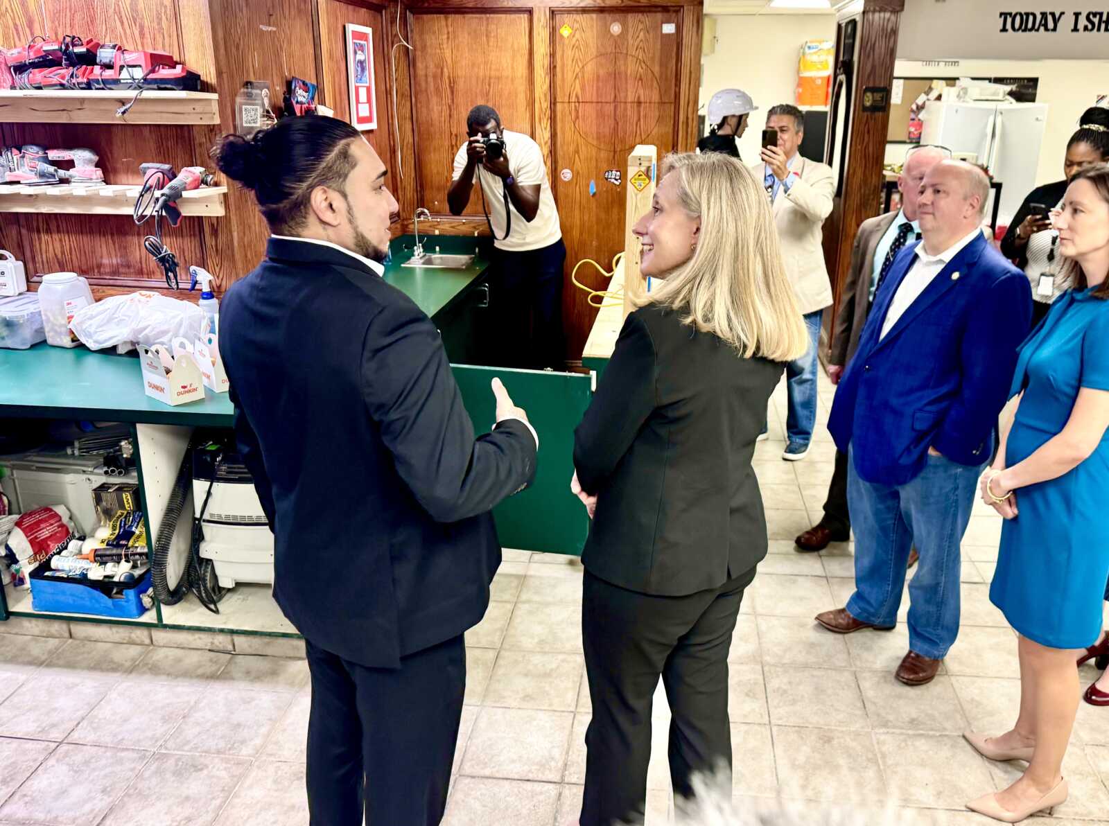 Man in a dark suit talks to a woman in a black business suit inside a shop while several people look on and photographers snap photos in the background.