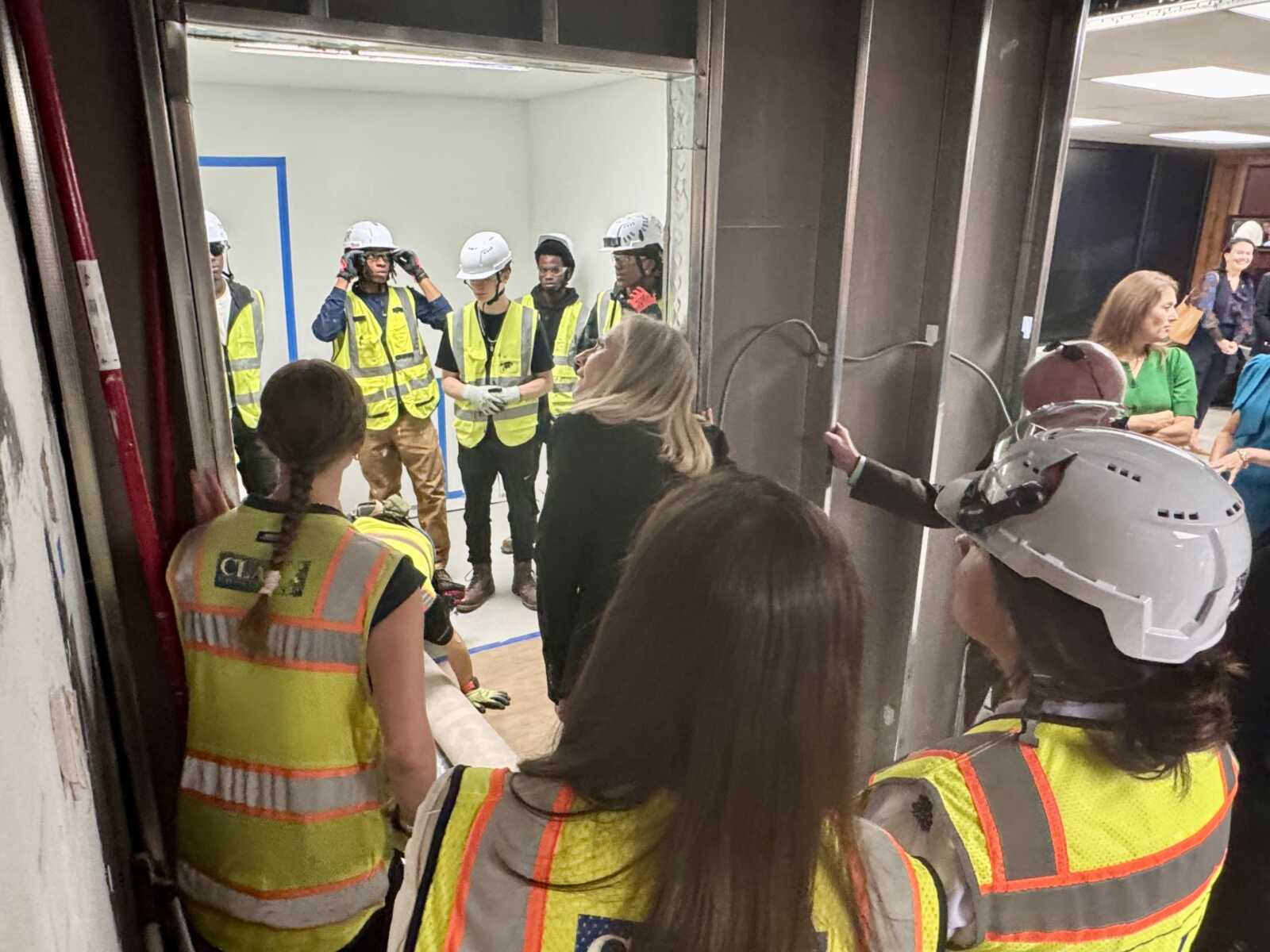 Group of construction workers in hard hats and high-visibility vests inside a building undergoing construction, with onlookers nearby.