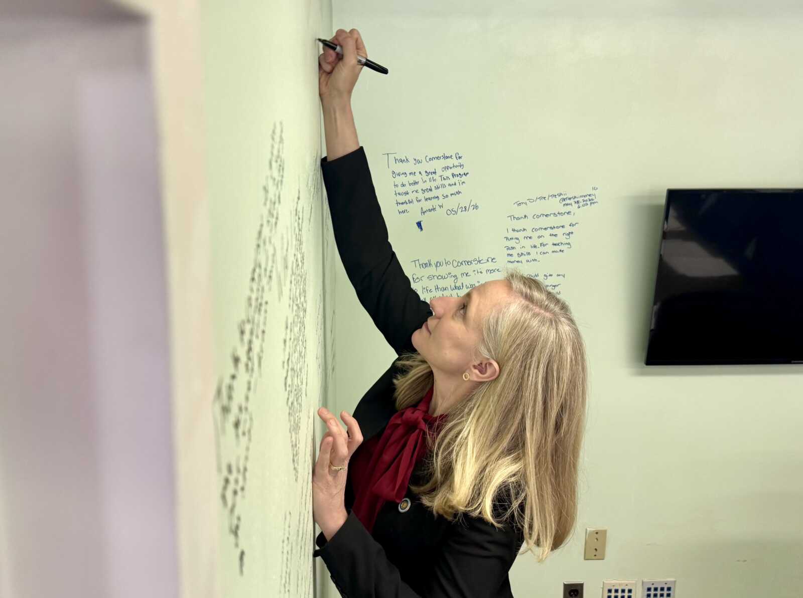 Woman in a blazer writes on a light-green wall with a marker; handwritten notes cover the wall beside a mounted TV.