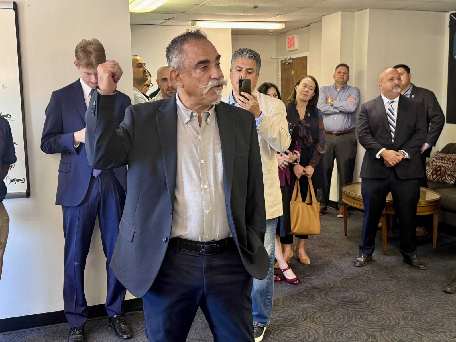 Older man in a dark suit raises his fist while speaking to a small group of professionals in an office hallway/conference area.