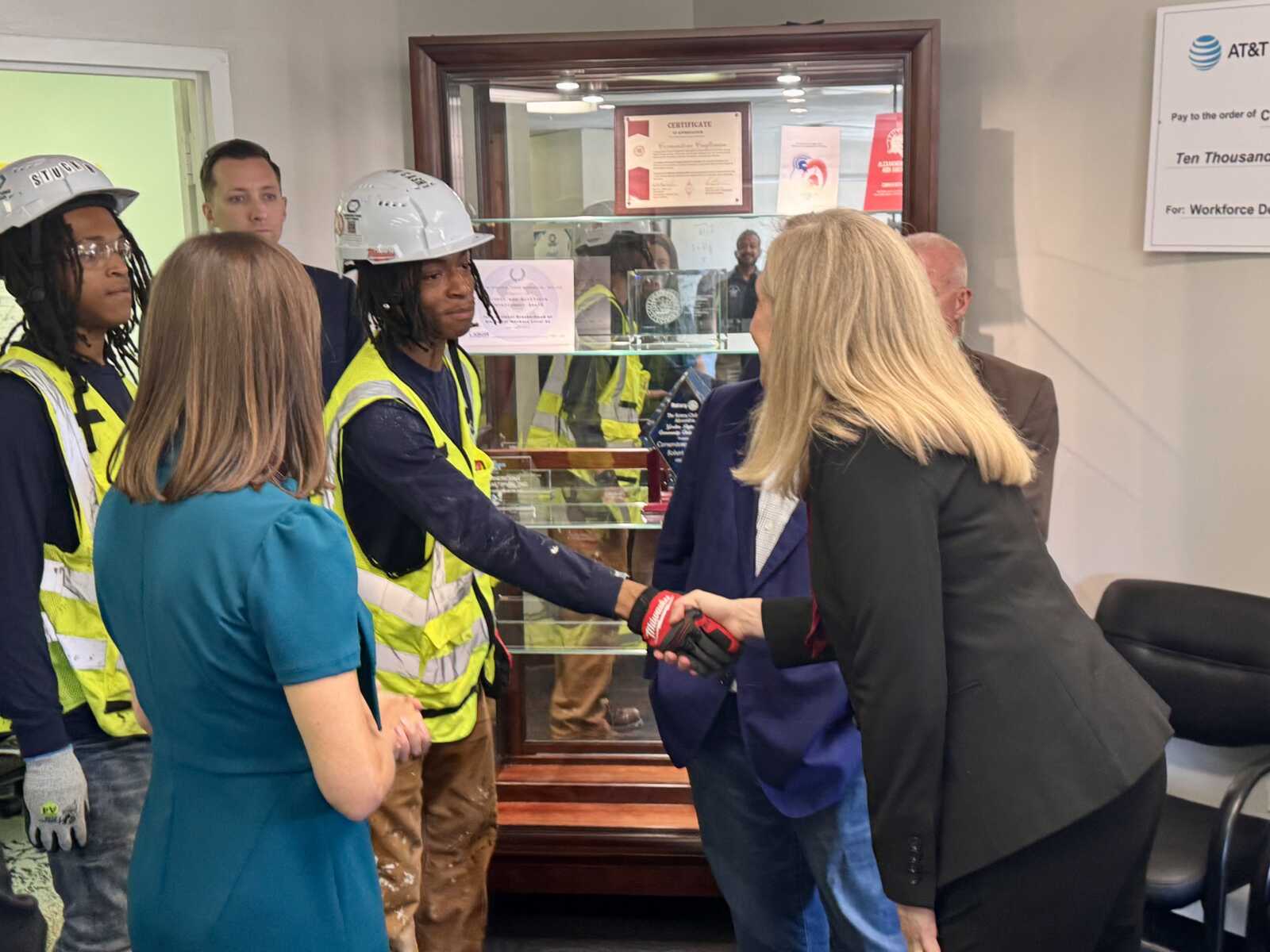 Businesswoman shakes hands with construction workers in a lobby, colleagues watching nearby.