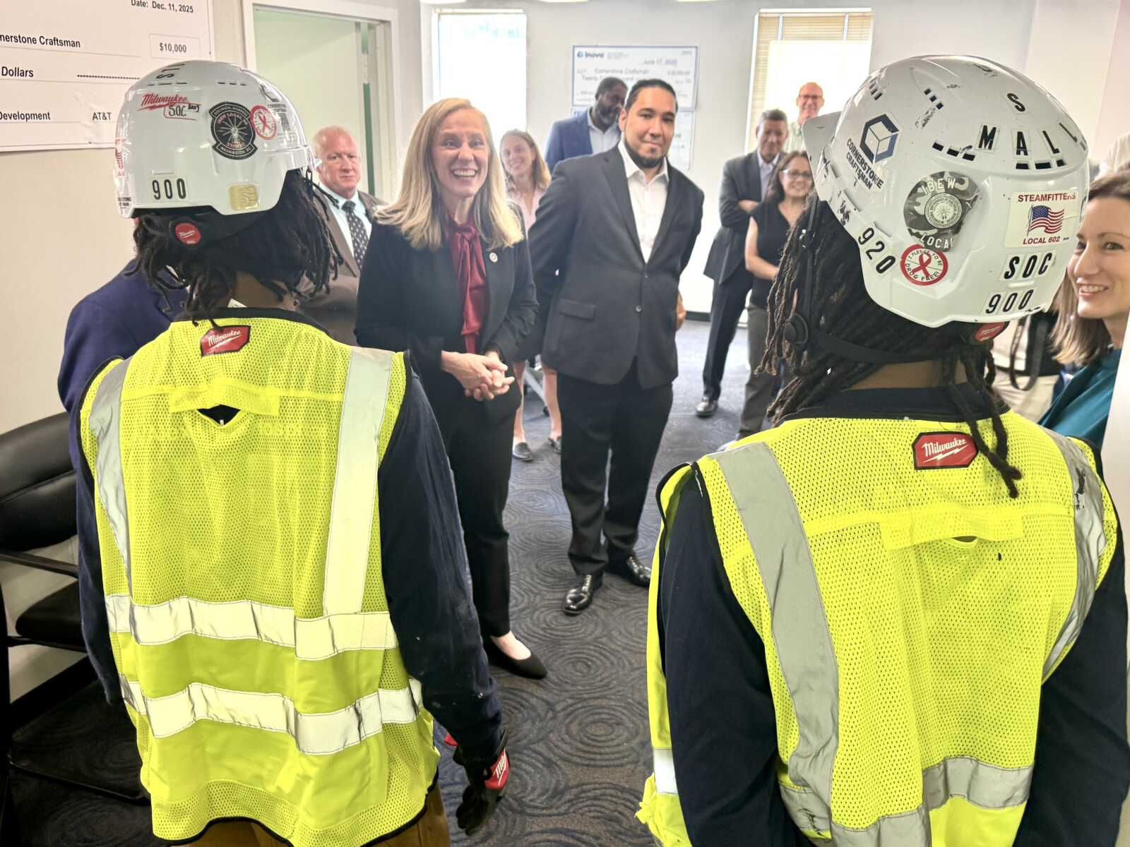 Two construction workers in high-visibility vests and white helmets greet a group of formally dressed professionals in an office lobby.