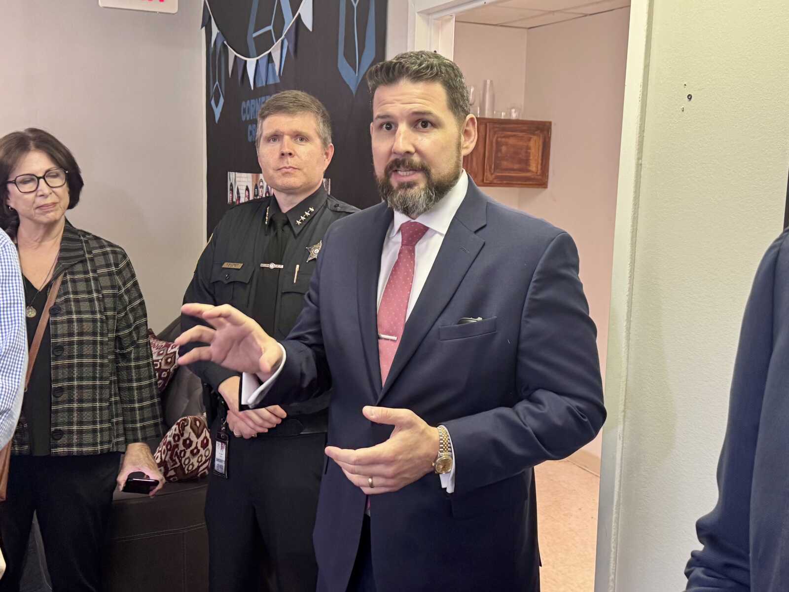 Man in a navy suit and pink tie speaks, gesturing with hands, with a uniformed officer and a woman listening nearby.
