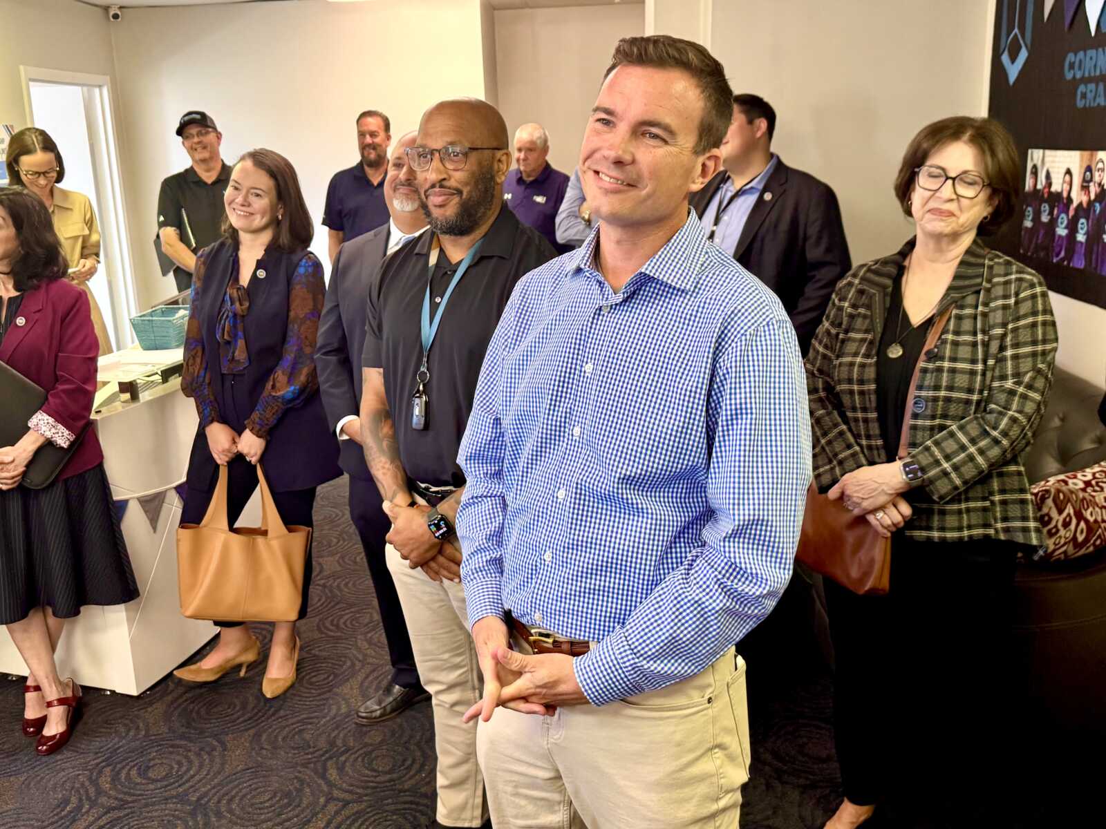 Group of professionally dressed people standing in a conference room, smiling at an event, with a man in a blue checkered shirt in the foreground.