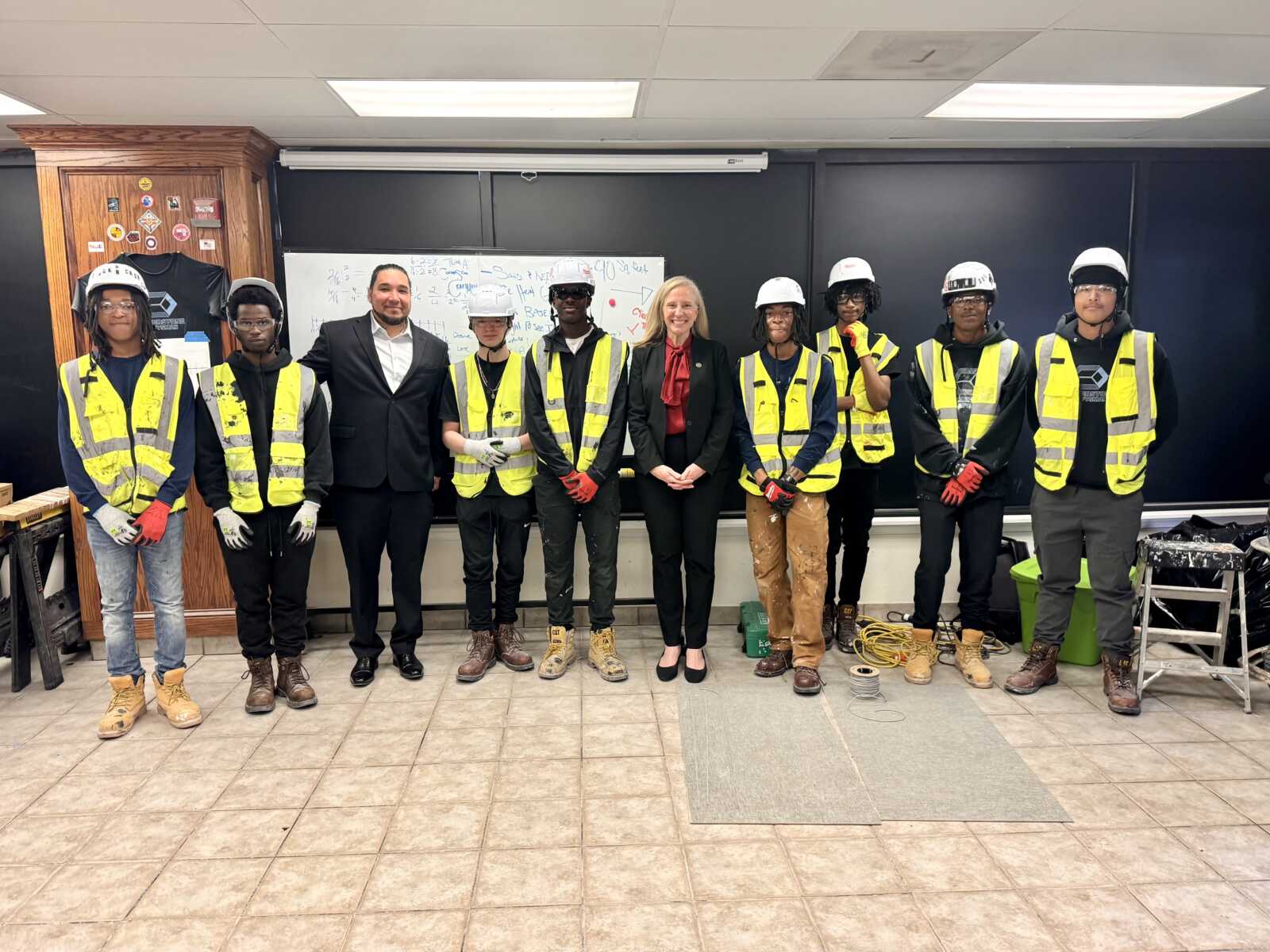 Ten students in yellow safety vests and hard hats stand with two adults in business attire for a group photo in a classroom workshop.