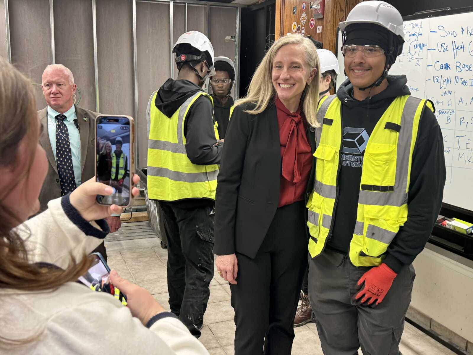 Woman in red blouse and black blazer posing with two construction workers in yellow safety vests and hard hats as others take photos nearby in an industrial space.