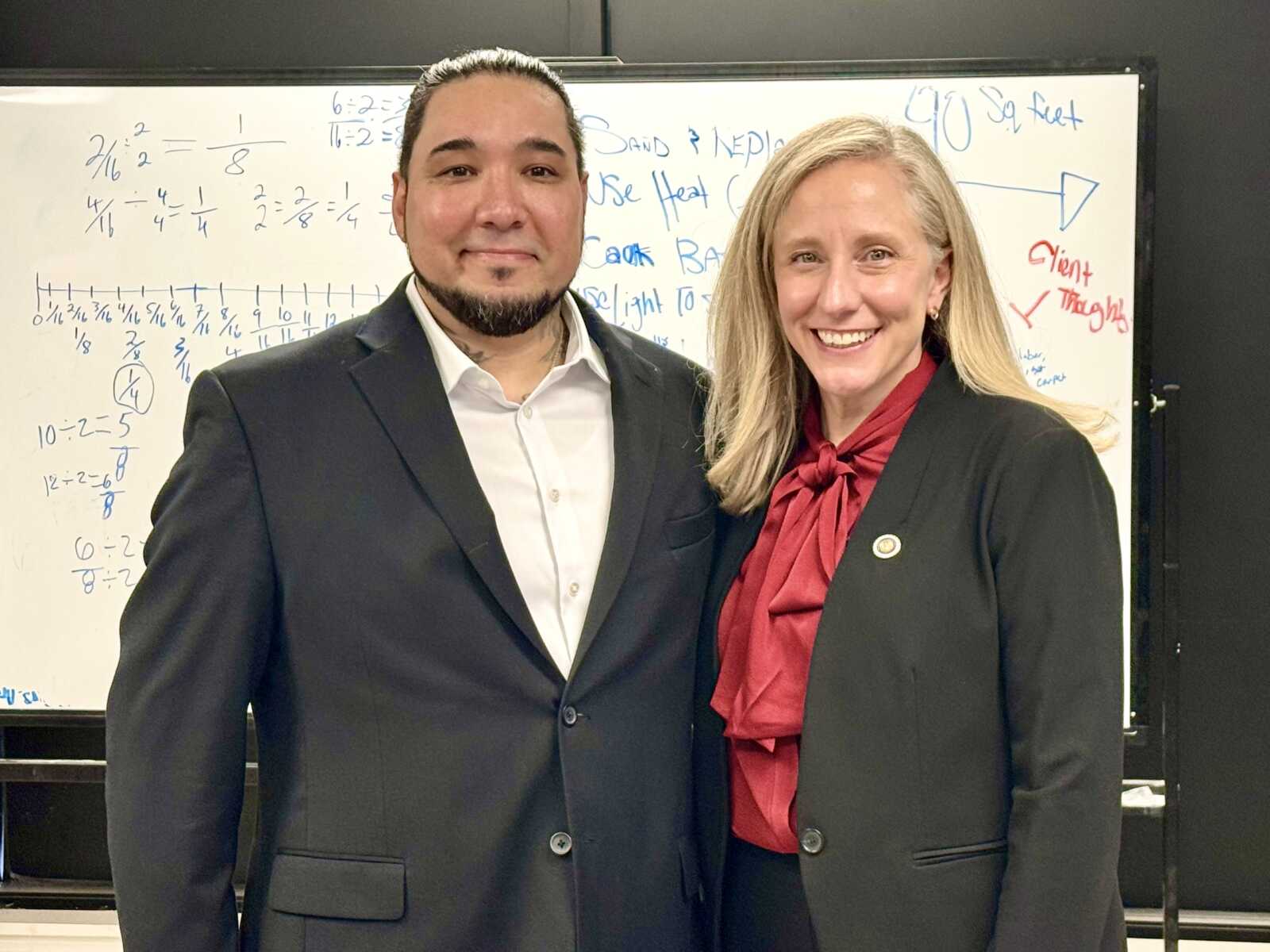 Two professionals in dark suits smile while posing in front of a whiteboard filled with handwritten equations and notes.