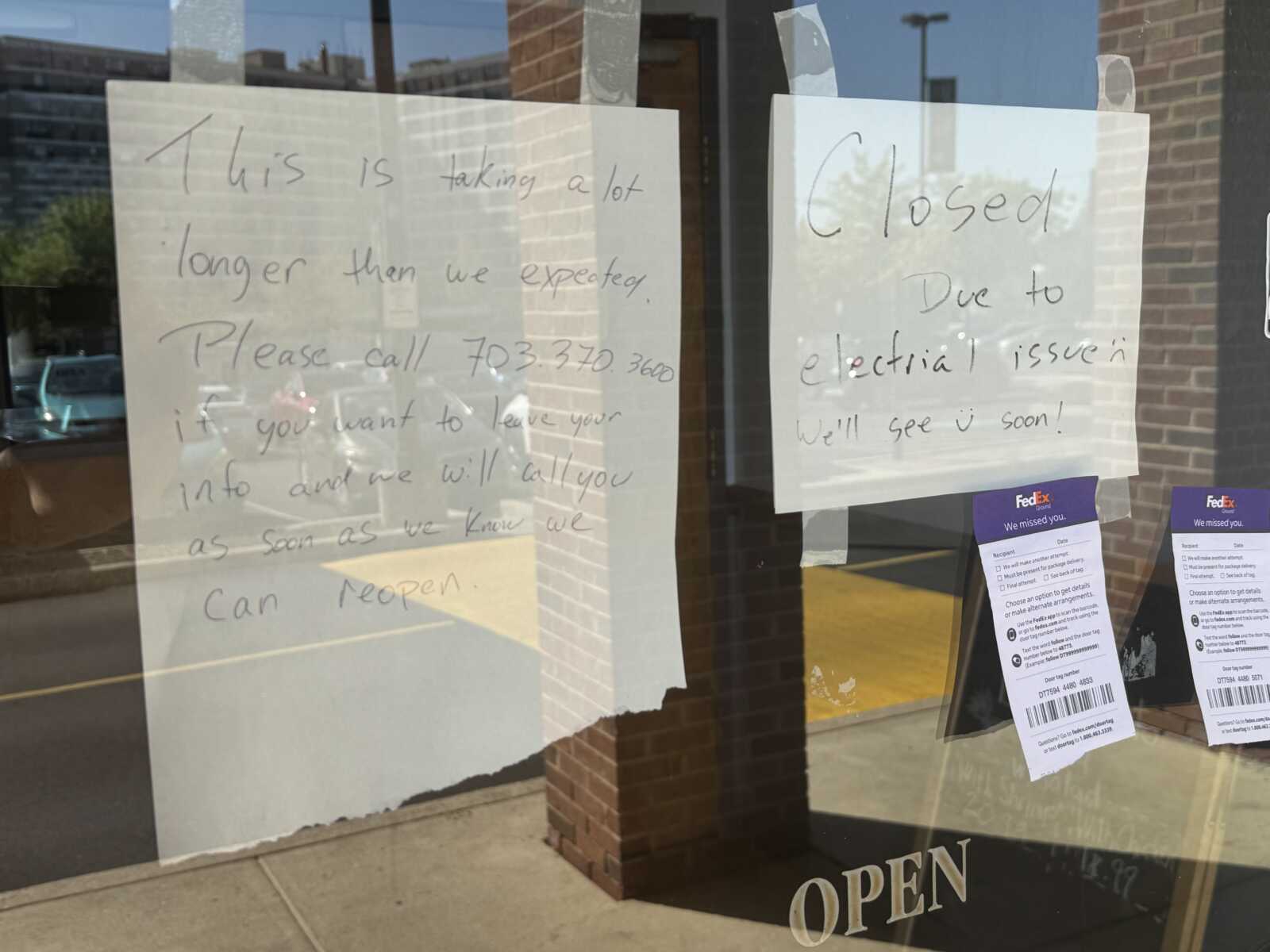 Storefront window with taped handwritten signs: one says 'CLOSED Due to electrical issue' and another notes 'We'll see you soon!', plus 'OPEN' at the bottom. Reflective glass shows street scene.