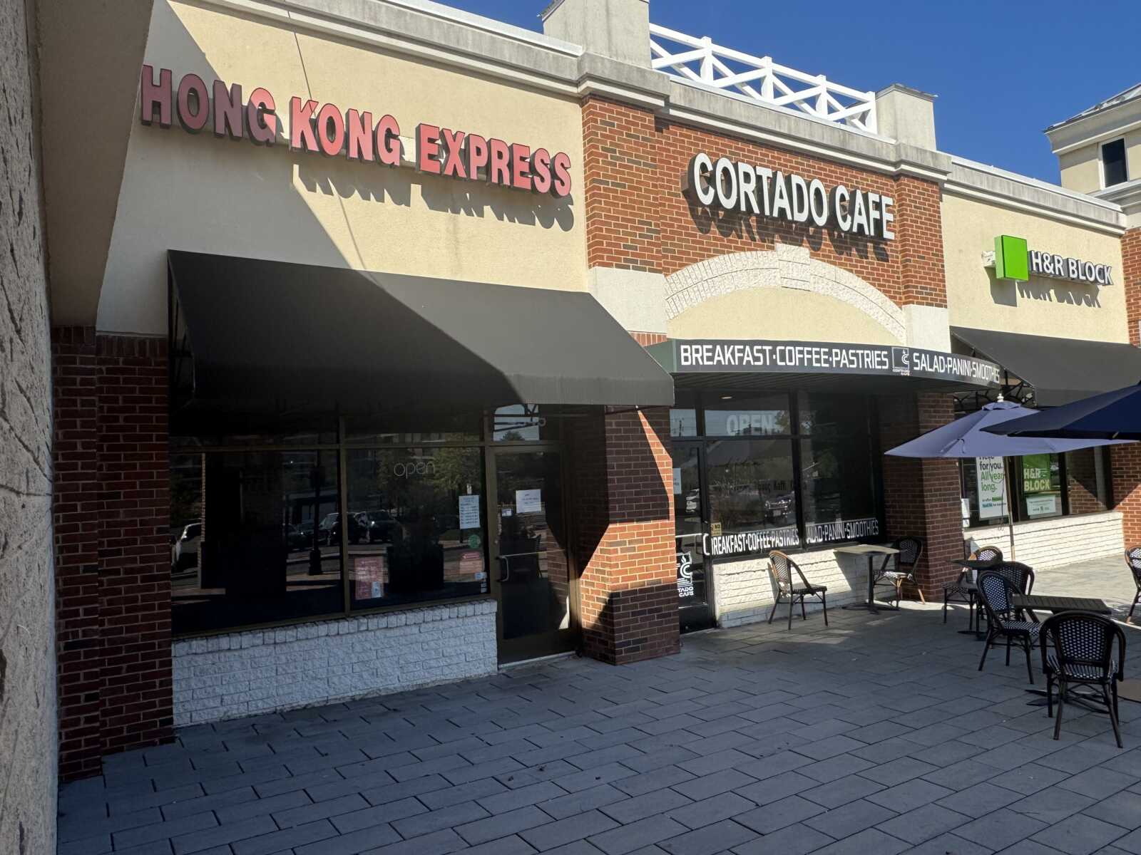 Exterior view of a café row with signs 'Hong Kong Express' and 'Cortado Cafe' on a brick storefront, with black awnings and outdoor seating under umbrellas.
