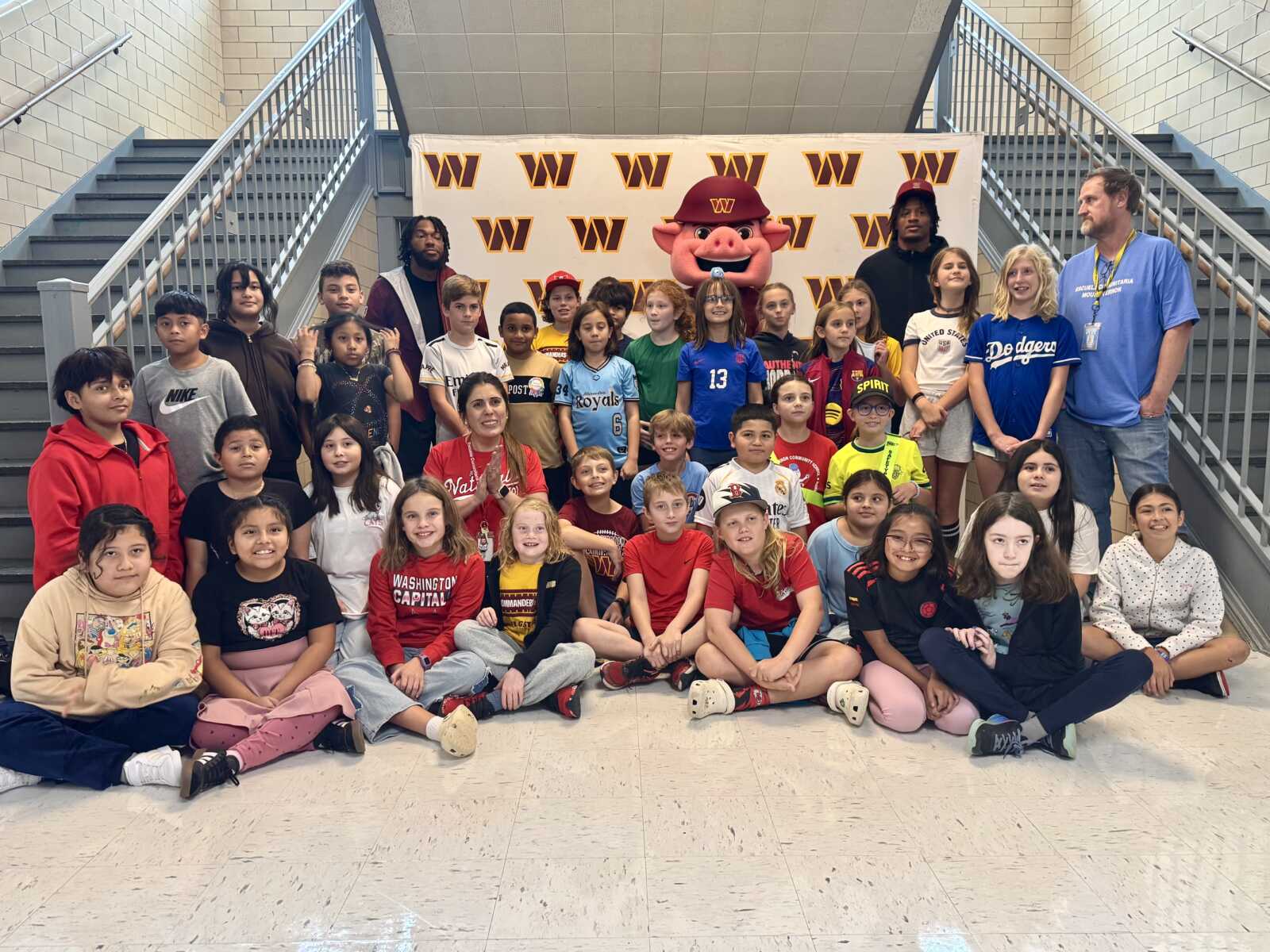 Large group of children posing for a school photo on stairs and floor, with a pig mascot in a red cap at the back and a Ws backdrop behind them.