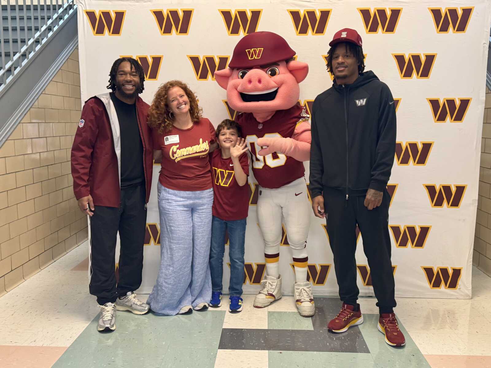Five people pose with a pig mascot in a burgundy football uniform in front of a W-pattern backdrop; smiling family/group photo in a school setting.
