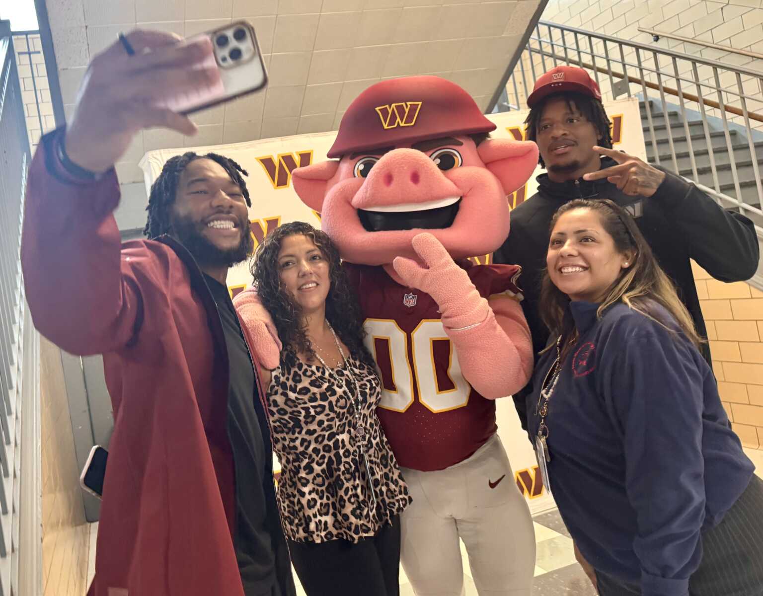 Four people posing with a pink pig mascot in a burgundy uniform, taking a selfie with a smartphone in a school hallway.