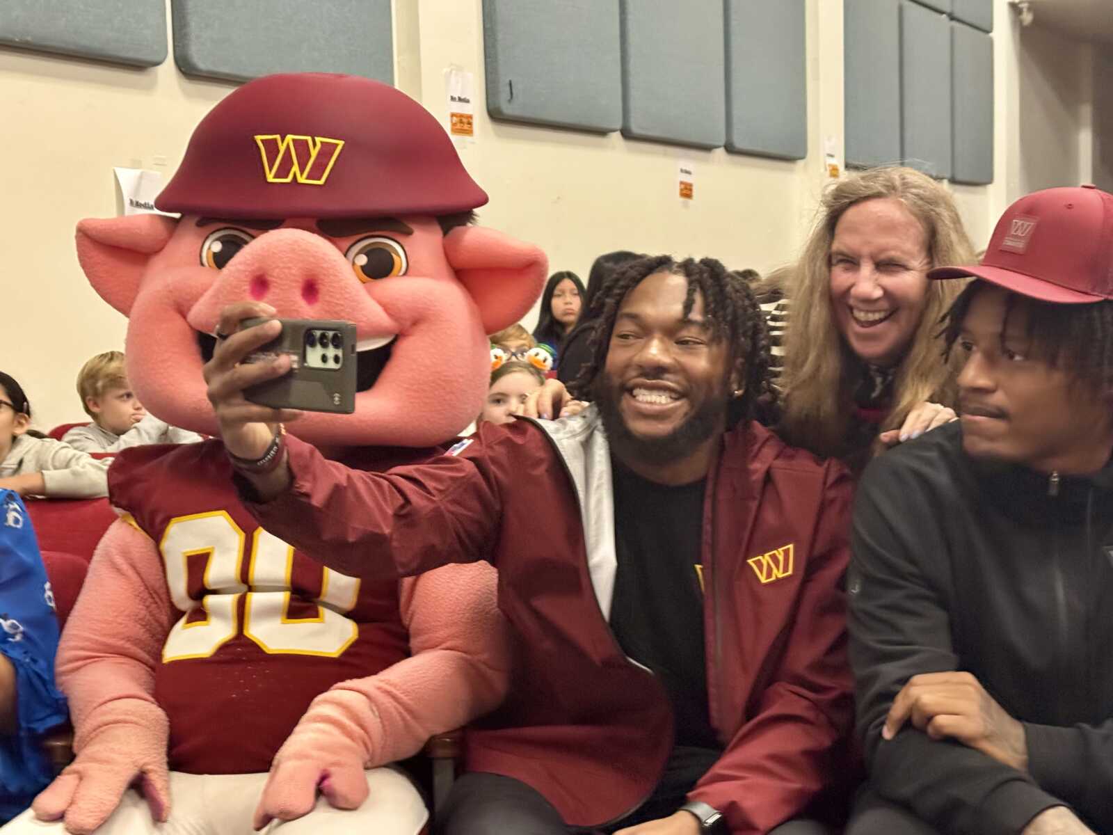 Person in a maroon mascot suit with a pig head takes a selfie with friends in a stadium setting; visible 'W' logos on hat and jacket and several smiling people around.