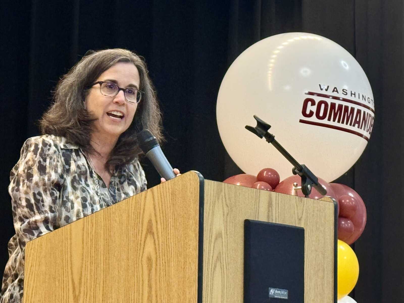 Woman in a patterned blouse speaks into a handheld microphone at a wooden podium, with balloons in the background on a stage buttoned up for an event.