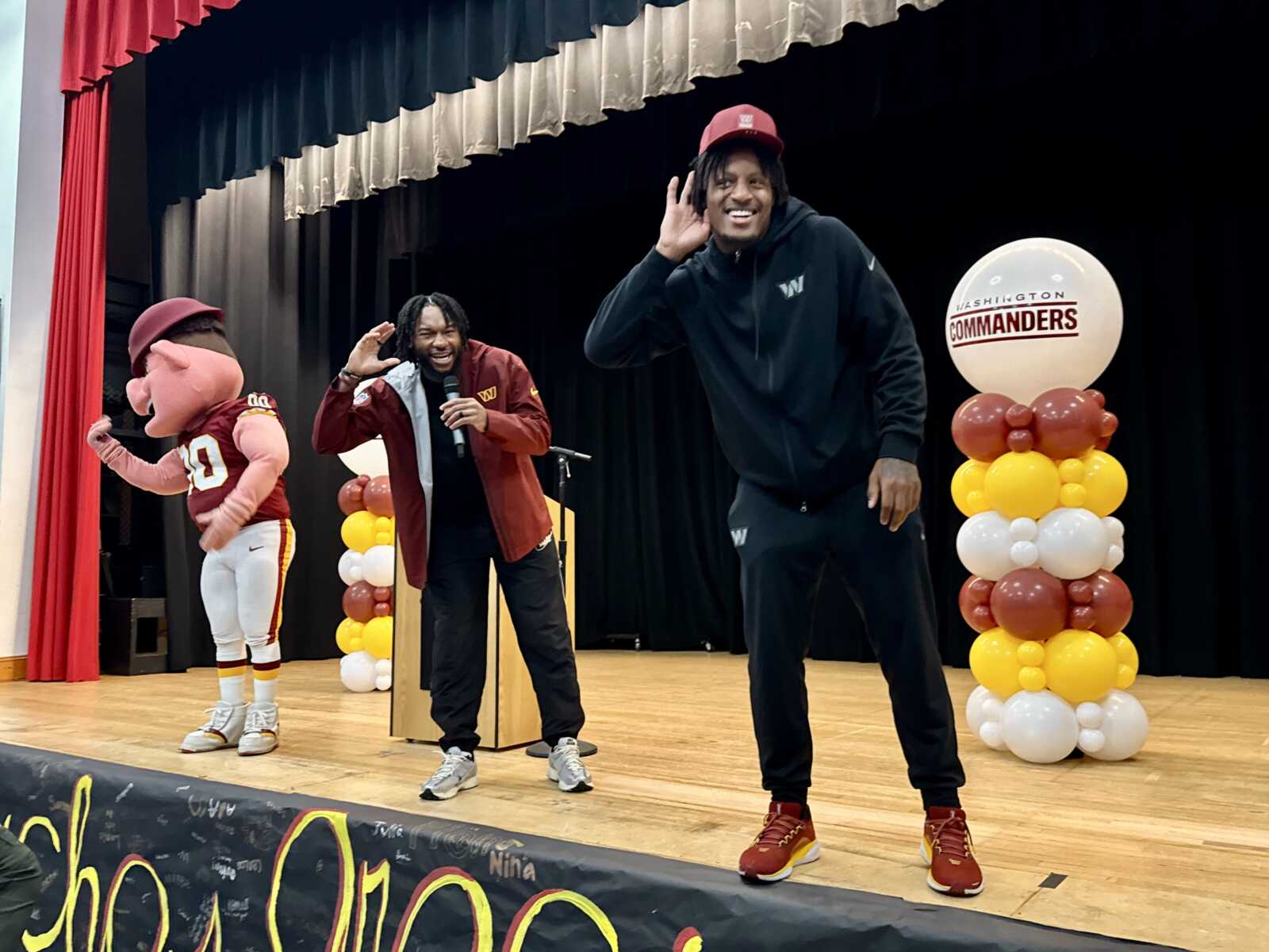 On a stage, a football mascot in burgundy and white stands left while a host with a microphone and a man in a black hoodie wave at the audience, with balloon columns and a Commanders logo in the background.