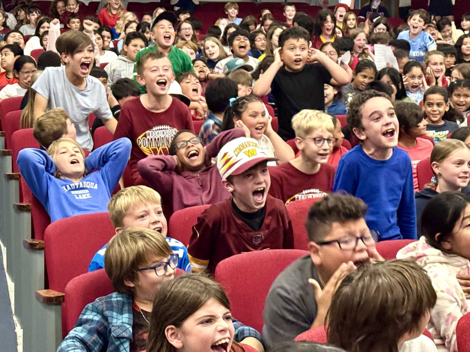 Crowded group of kids sitting in red theater seats, cheering and shouting with wide smiles.