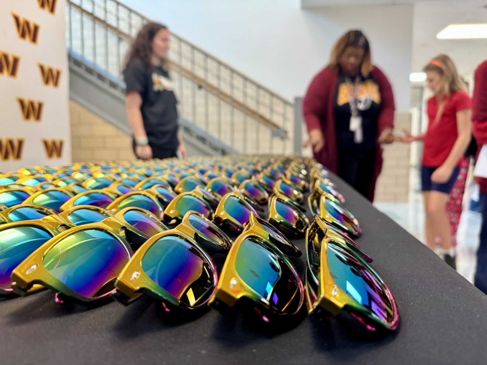 Row of rainbow-reflective sunglasses laid out on a black table, with people chatting in the background at a school-like event.
